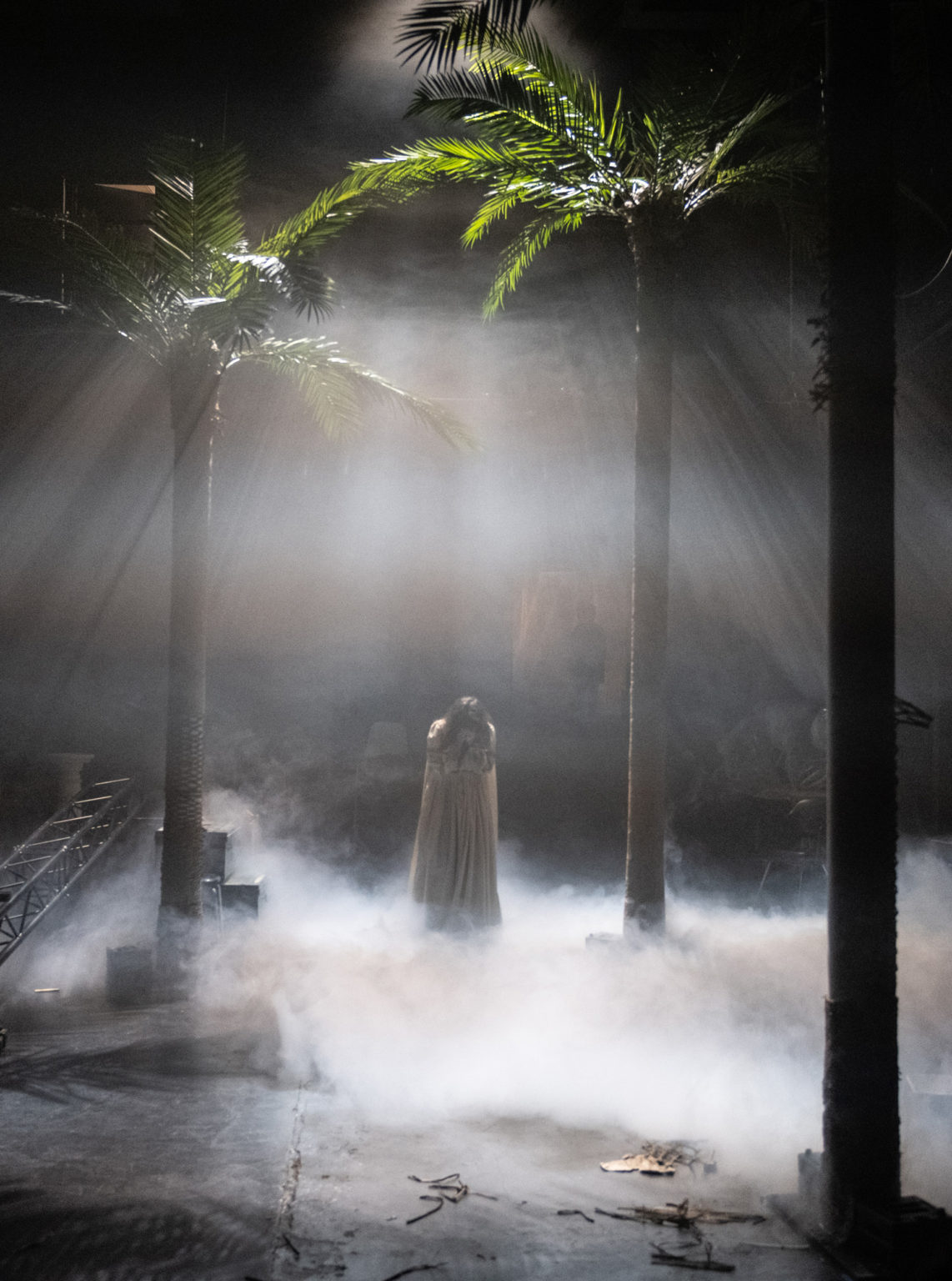 Photo du spectacle "Paradis fin de règne" où l'on voit la comédienne Sarah Espour, au milieu du plateau, dans un halo de lumière et de fumée, le visage enfouie dans ses mains.