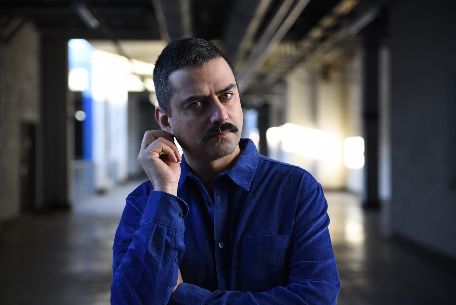 Portrait de l'artiste Gurshad Shaheman qui se touche l'oreille. Il porte une chemise bleue.