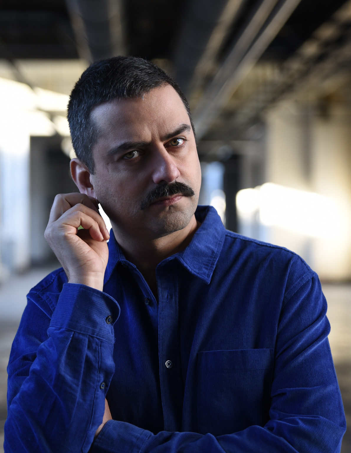 Portrait de l'artiste Gurshad Shaheman qui se touche l'oreille. Il porte une chemise bleue.