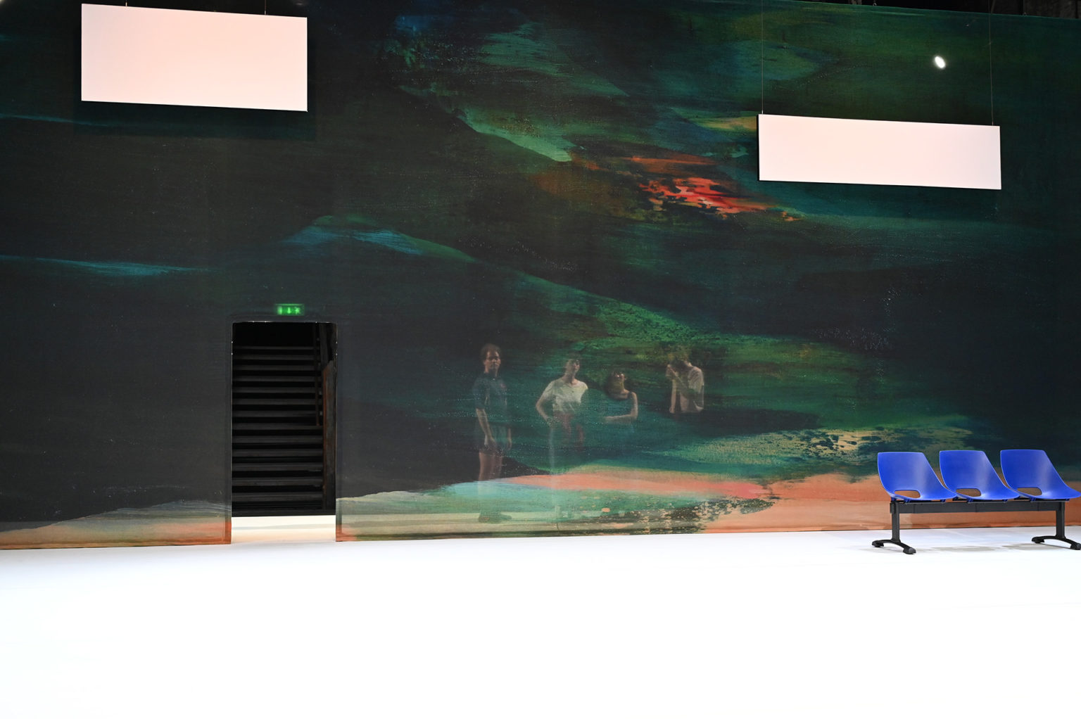 Les quatre acteurs et actrices sont derrière un mur de couleur transparent et regardent vers nous. Il y a un banc bleu.