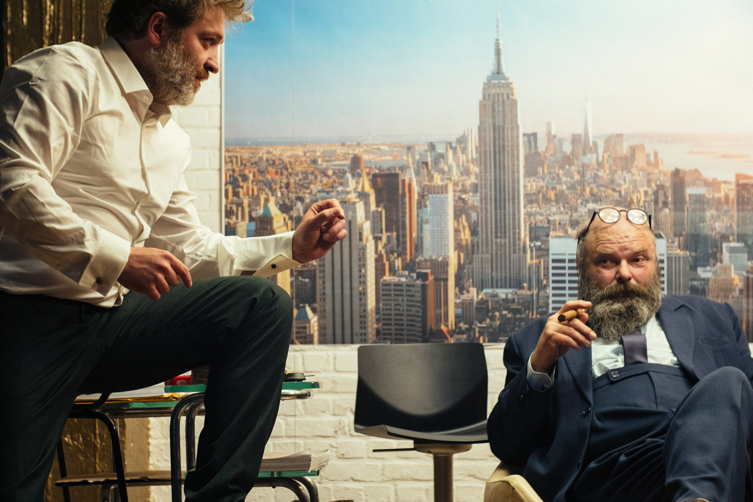 Les acteurs Bert Haelvoet et Damiaan De Schrijver discutent, cigares à la main. Derrière eux, une photo de New York où l'on voit l'Empire State Building.
