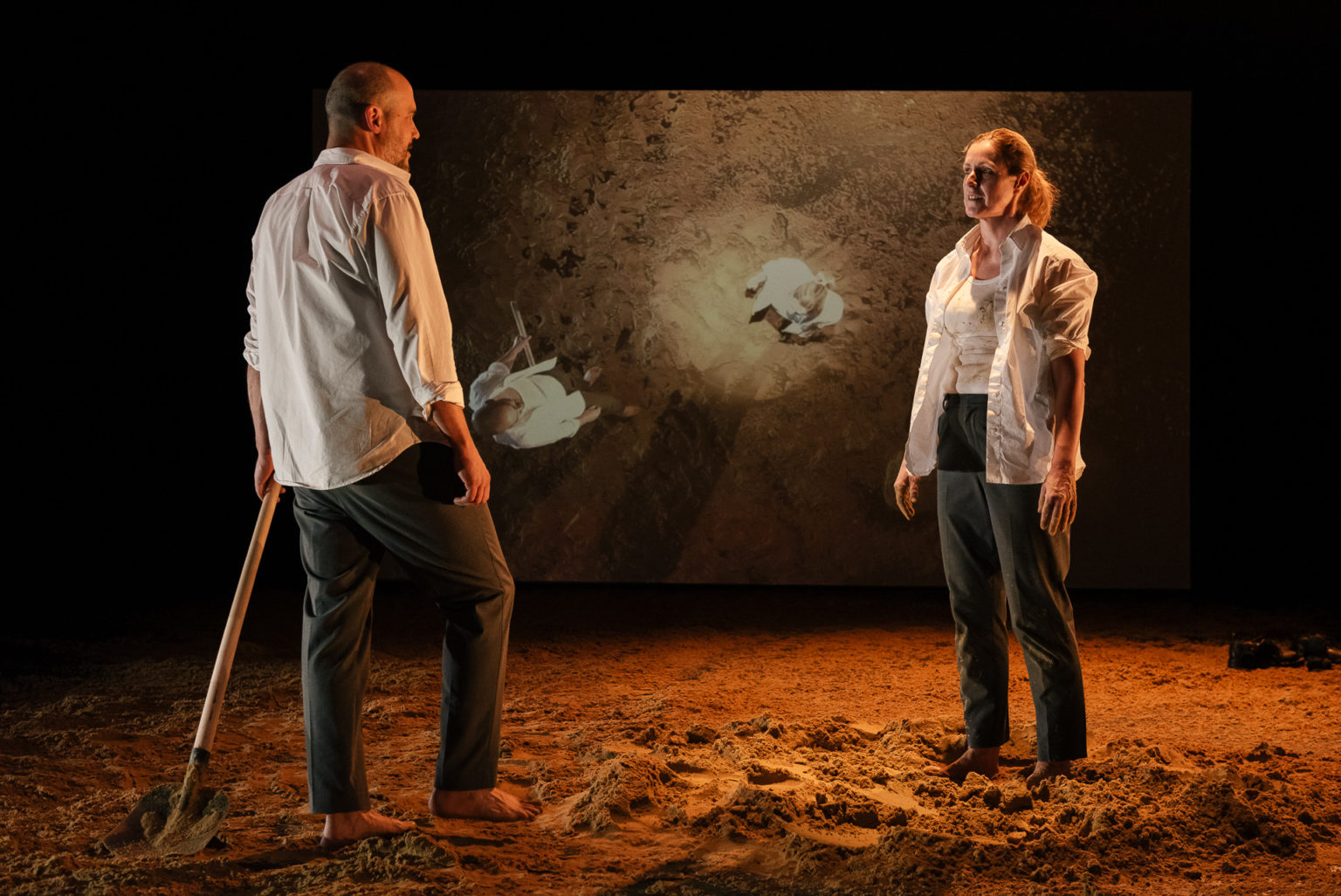 Rachid Benbouchta et Geneviève Damas se font face. Il tient une pelle en main. Derrière eux, un écran projette leur image. Du sable recouvre le plateau.
