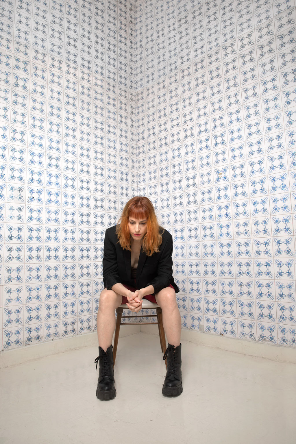 Marina Otero est assise sur une chaise. Elle est habillée en noir. Elle regarde le sol. Derrière elle, un mur de carreaux blancs et bleus.