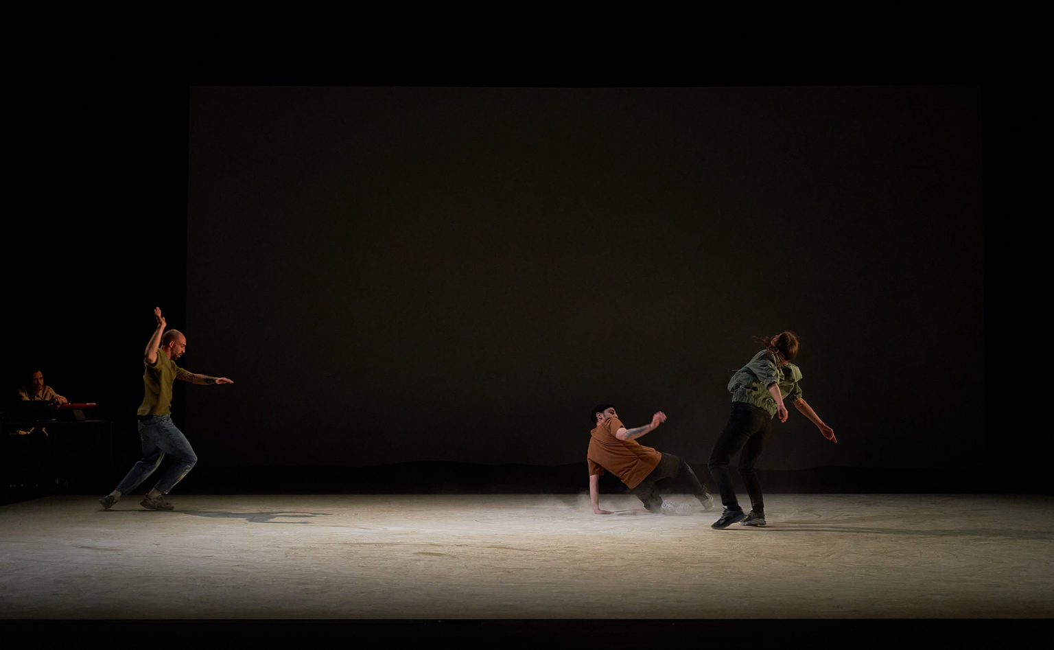 Photo du spectacle "Paysage" où l'on voit les 3 danseurs au milieu du grand plateau vide. Le sol est recouvert d'une poussière. L'un danse au sol.