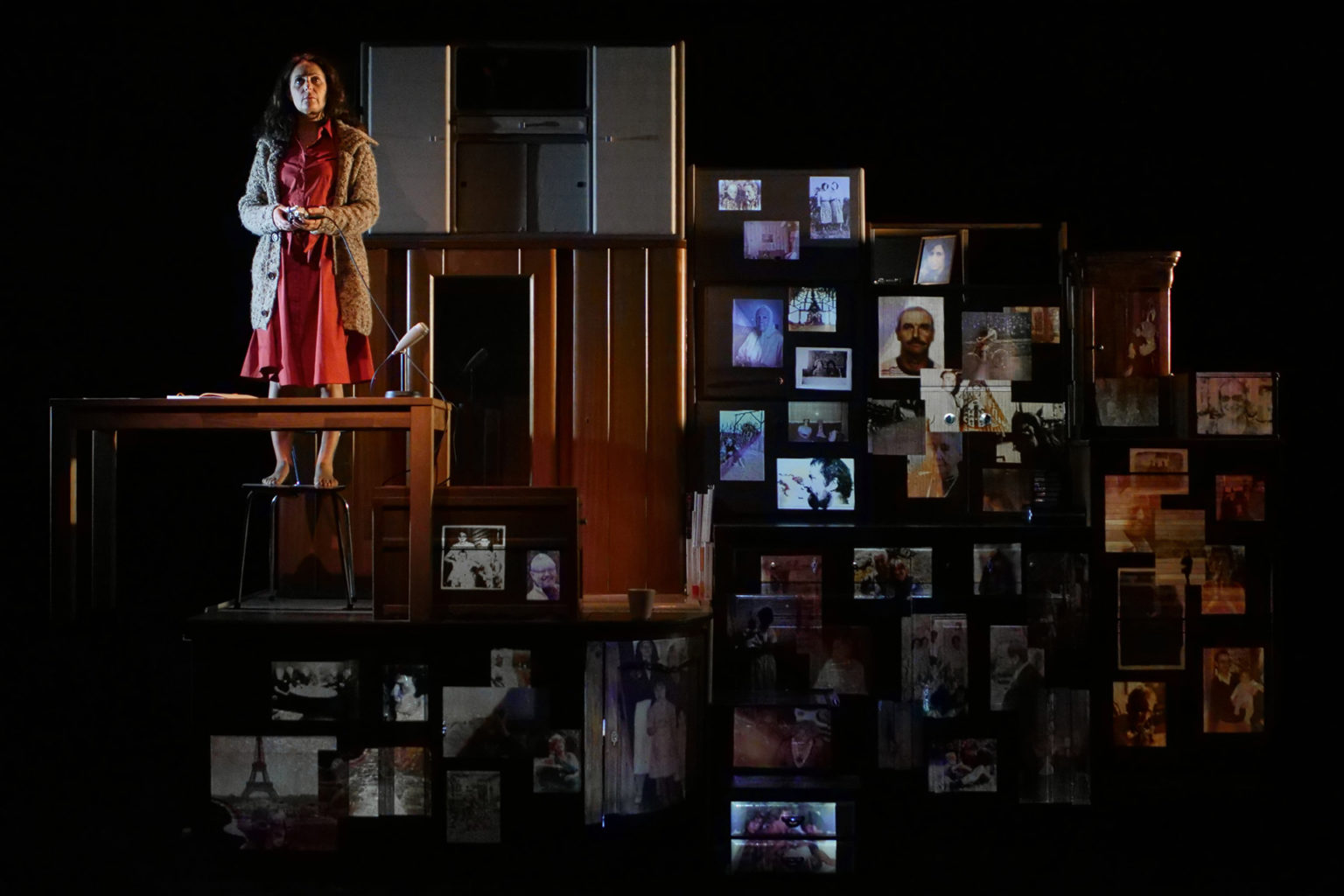 Jasmina Douieb est debout, sur un tabouret. Elle porte une robe rouge et un gilet. Elle est devant un grand meuble à tiroirs. Elle tient un enregistreur en mains.