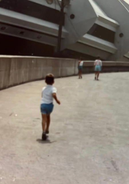 Photo de famille où l'on voit un petit garçon - Dany Boudreault - courir devant un grand bâtiment en béton qui ressemble à un vaisseau spatial.