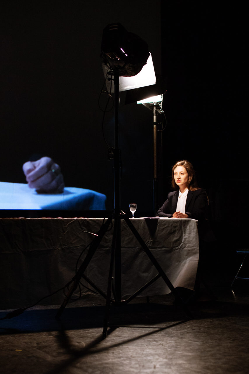Ariane Loze est assise à une table, comme sur un plateau de cinéma. Un écran est derrière elle et projette son image.