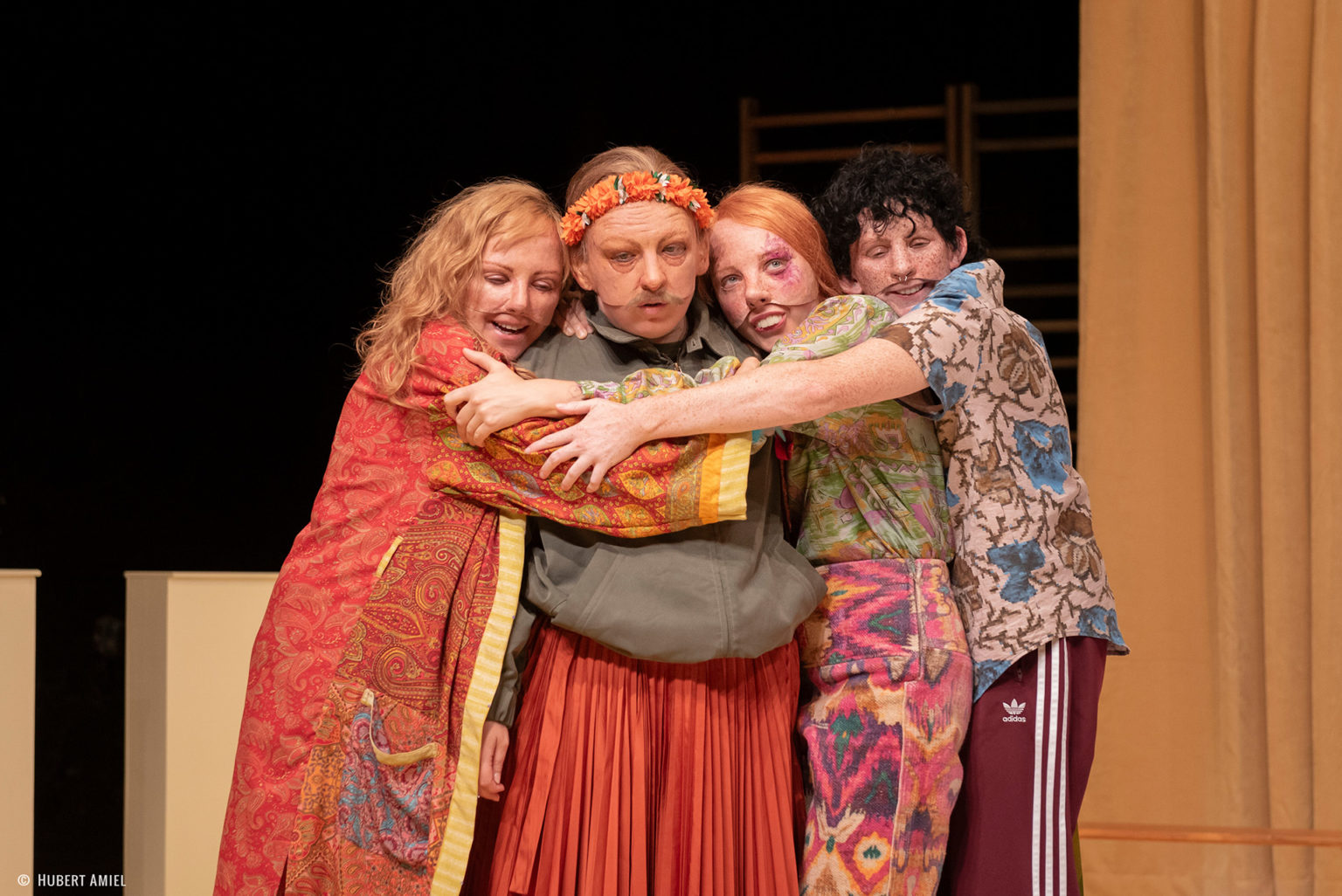 Photo du spectacle L'amour c'est pour du beurre où l'on voit 4 des comédiennes qui se font un câlin. Elles portent des masques réalistes et des vêtements colorés.