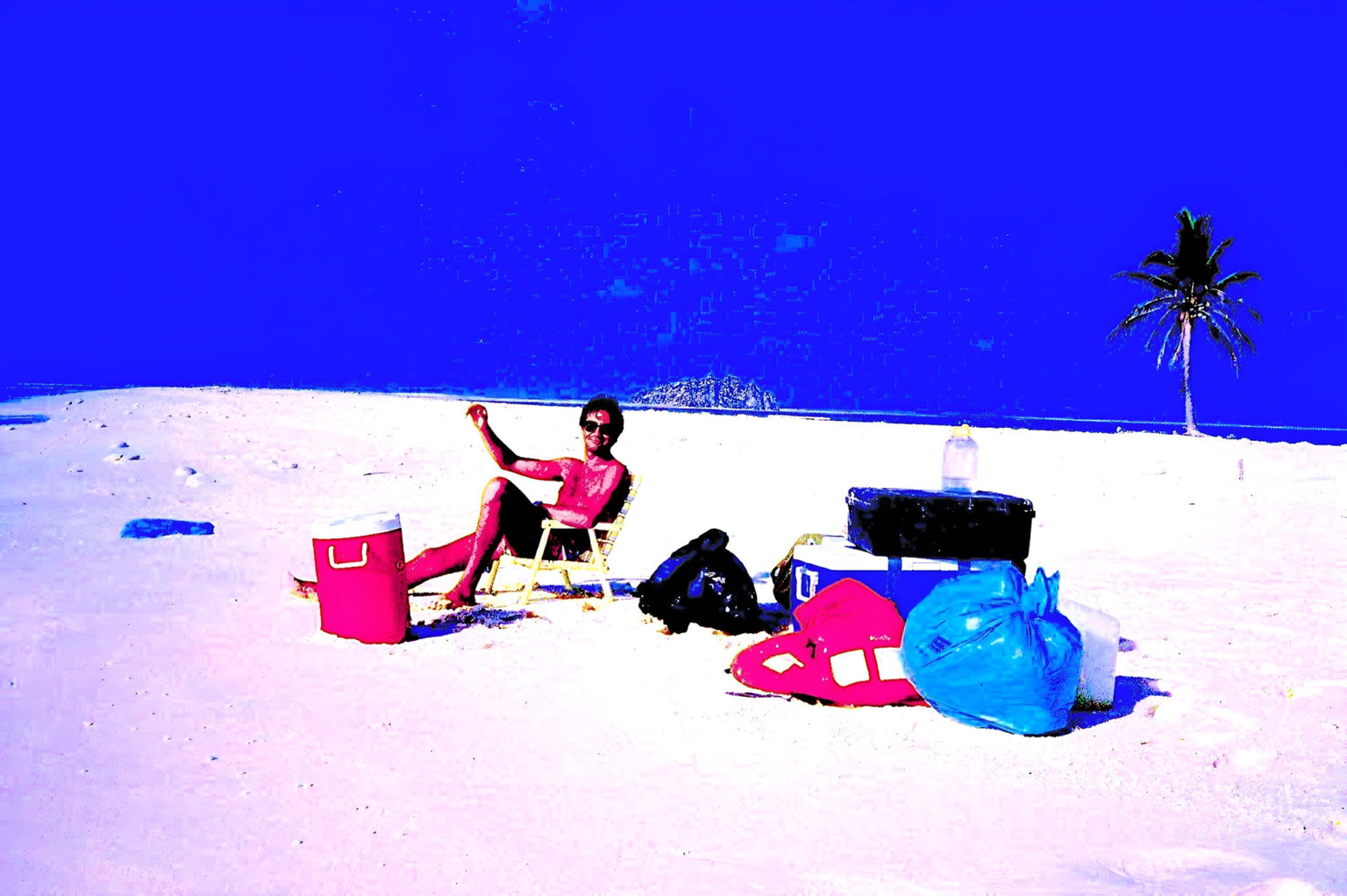 Un homme en maillot de bain est assis sur une chaise de plage sur le sable d'une plage avec, autour de lui des affaires de voyage. Les couleurs dominantes sont le bleu, le rouge, le noir et le blanc. Les couleurs sont très saturées.