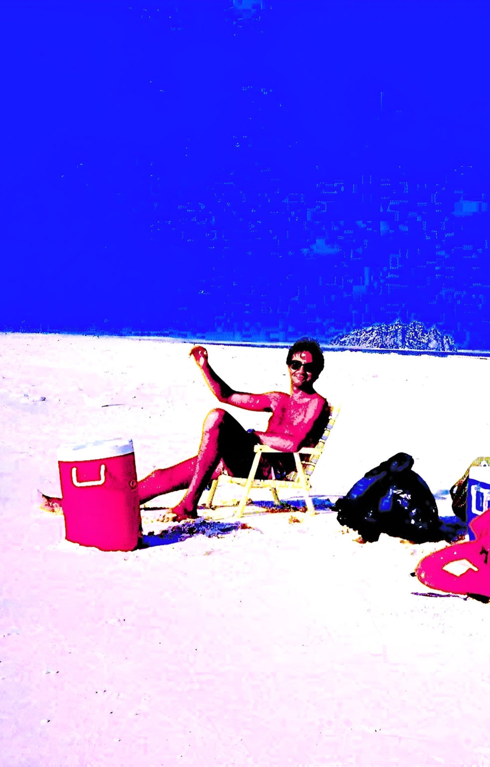 Un homme en maillot de bain est assis sur une chaise de plage sur le sable d'une plage avec, autour de lui des affaires de voyage. Les couleurs dominantes sont le bleu, le rouge, le noir et le blanc. Les couleurs sont très saturées.