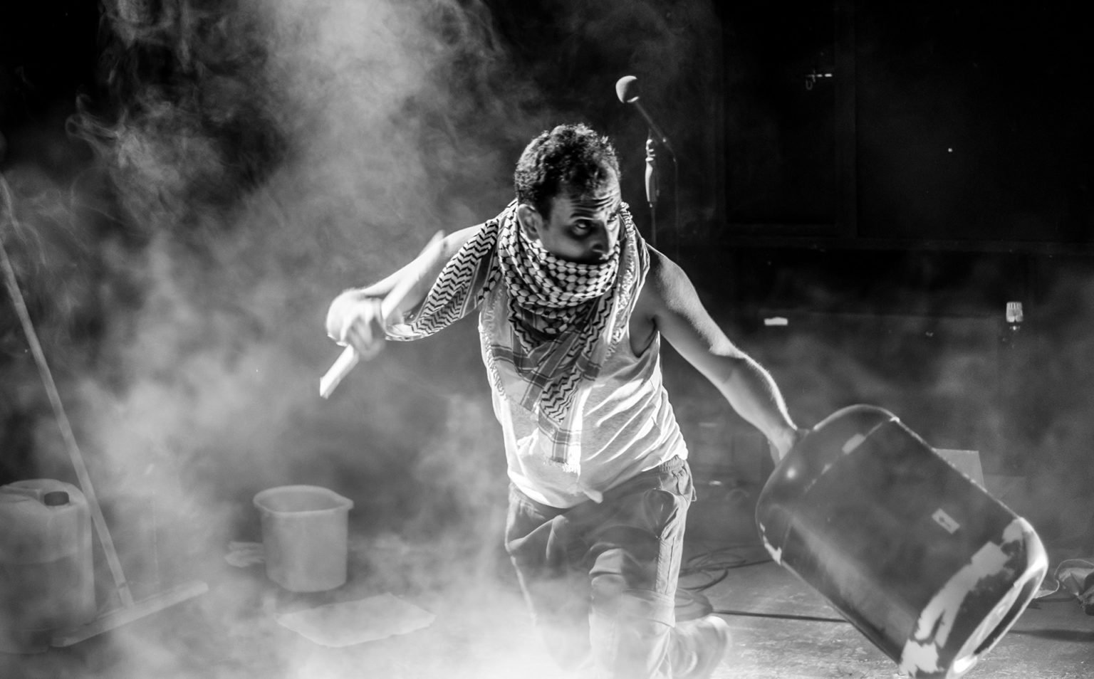 Photo en noir et blanc d'Ahmed Tobasi sur scène entrain de performer. Il porte un débardeur blanc et un   keffieh (écharpe à carreaux noir et blanc) couvre son cou et sa bouche.