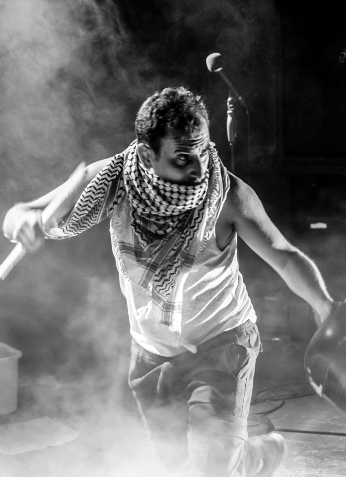 Photo en noir et blanc d'Ahmed Tobasi sur scène entrain de performer. Il porte un débardeur blanc et un keffieh (écharpe à carreaux noir et blanc) couvre son cou et sa bouche.