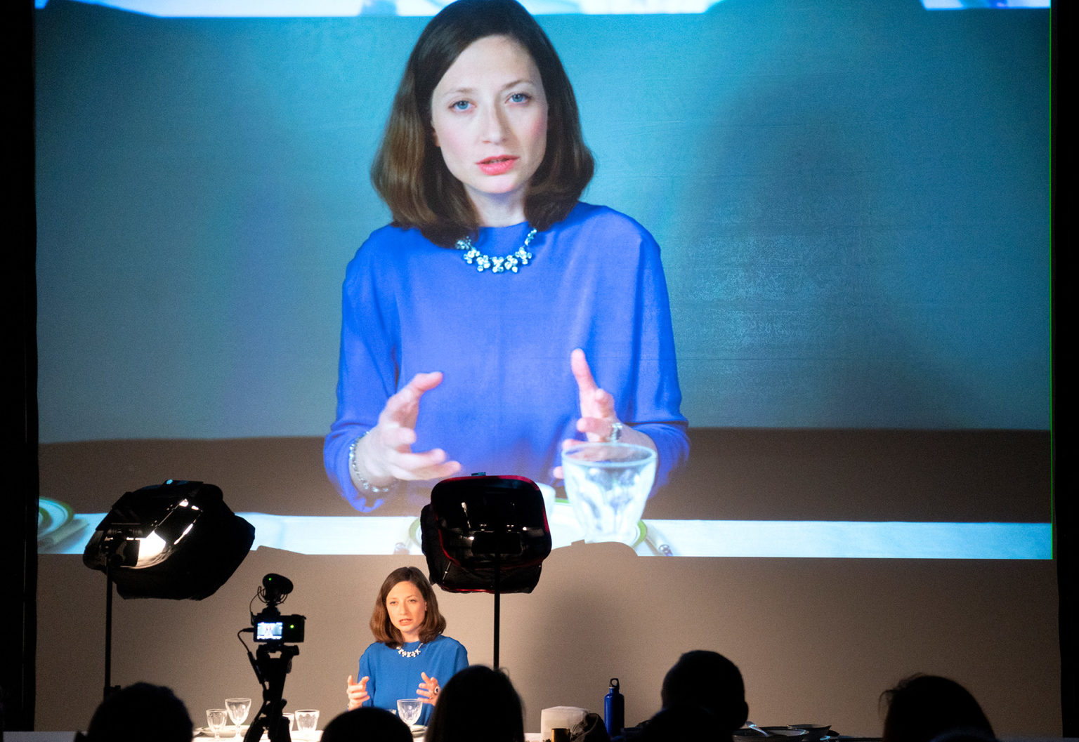 Ariane Loze en pleine performance, devant une grande table. Son image est projetée sur un écran géant. Elle porte un haut bleu.