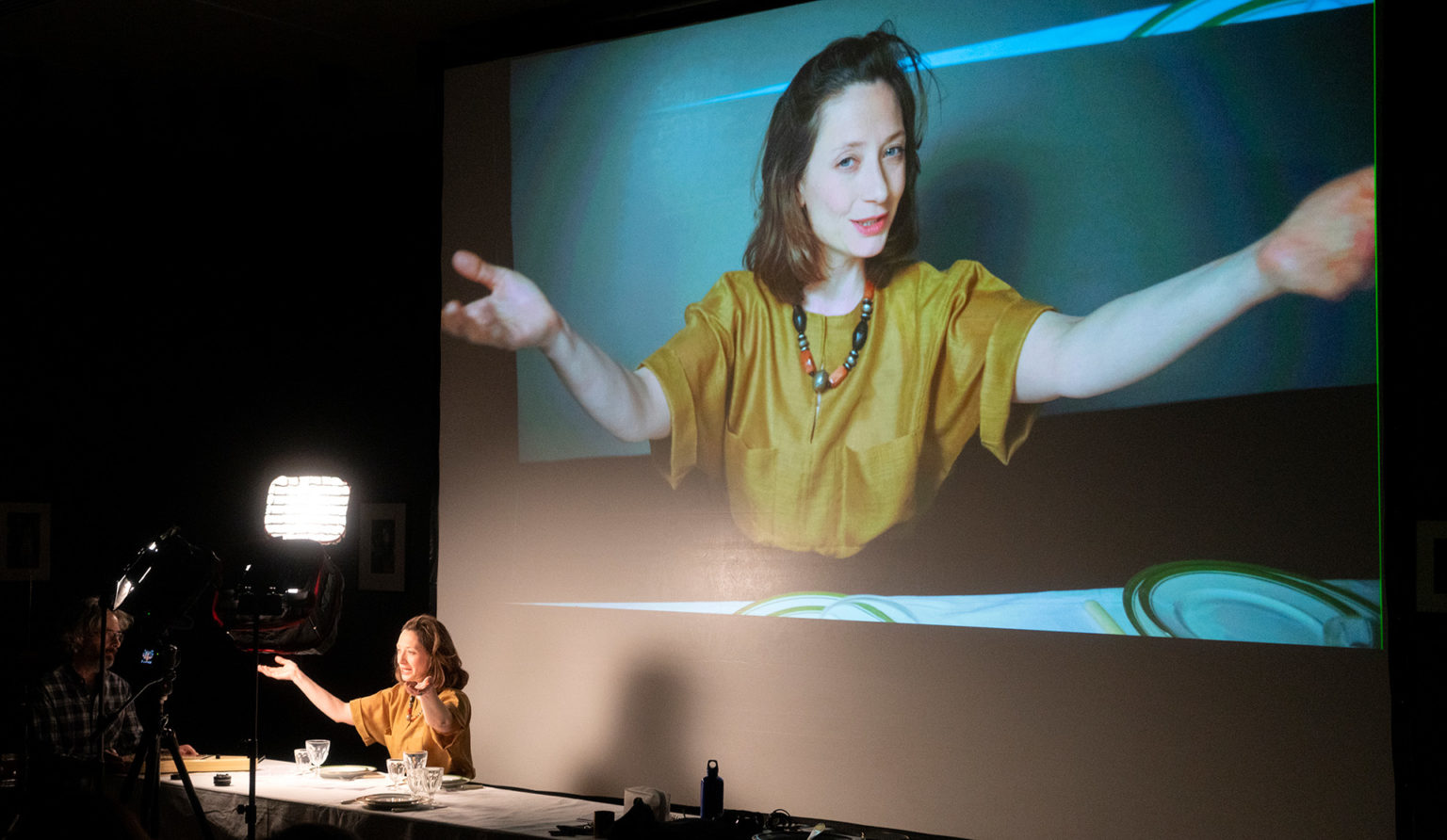 Ariane Loze en pleine performance, devant une grande table. Son image est projetée sur un écran géant. Elle porte un haut jaune.