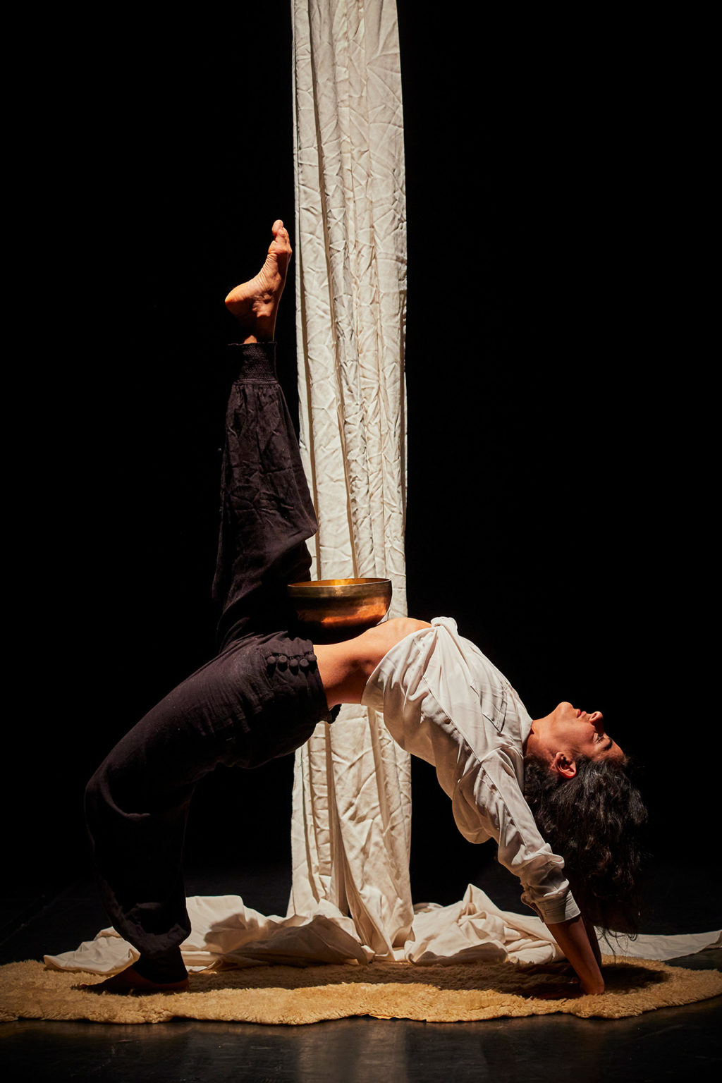 Photo d'Ashtar sur scène entrain de performer au pied d'un grand tissu suspendu. Elle porte un haut de couleur blanc similaire à la couleur du tissu. Et elle porte un pantalon noir.