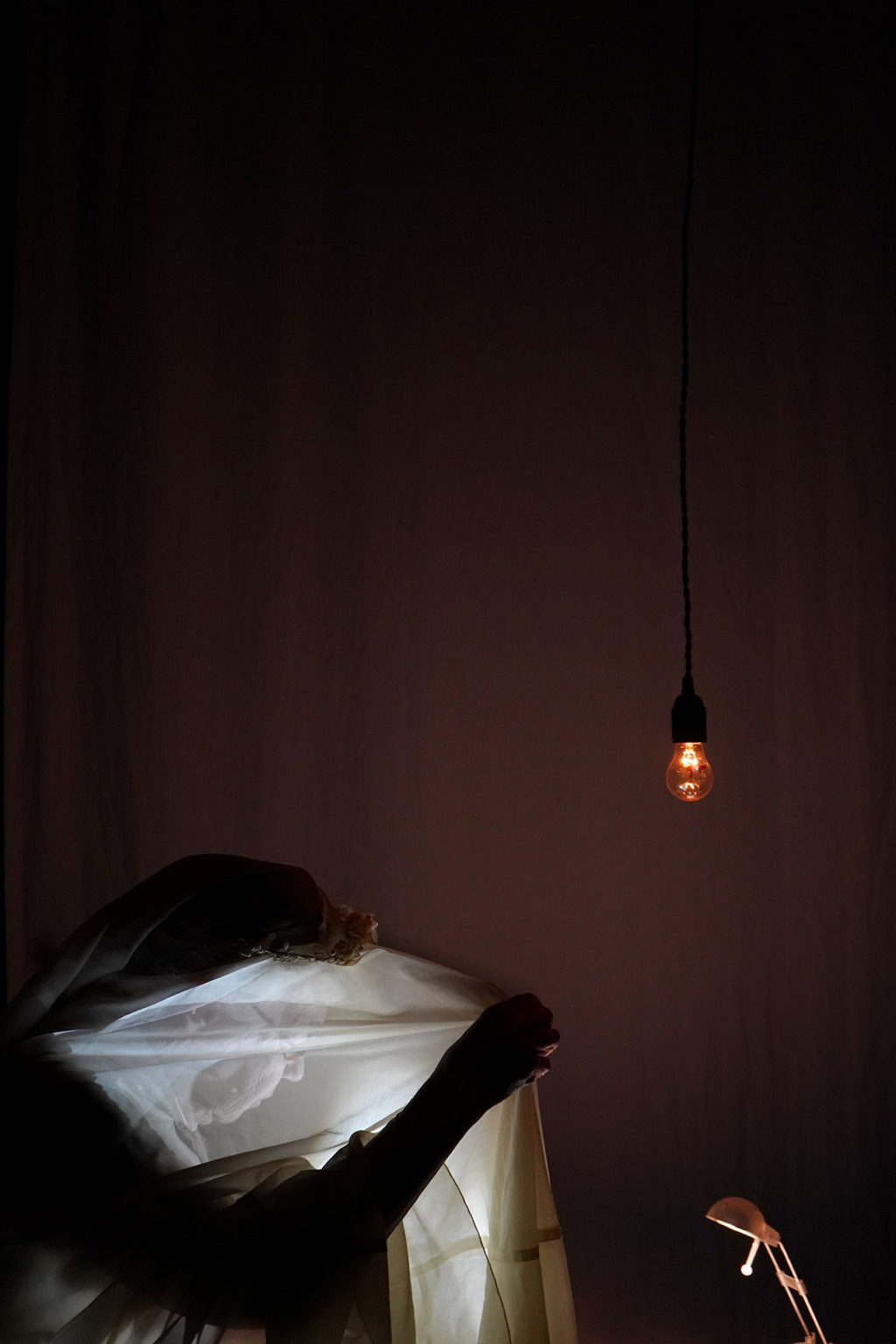 Photo d'un homme dans le noir qui est couvert par un tissu transparent blanc laissant apercevoir son profil. Sur le côté droit, une ampoule suspendue à un fil électrique et une lampe de chevet.