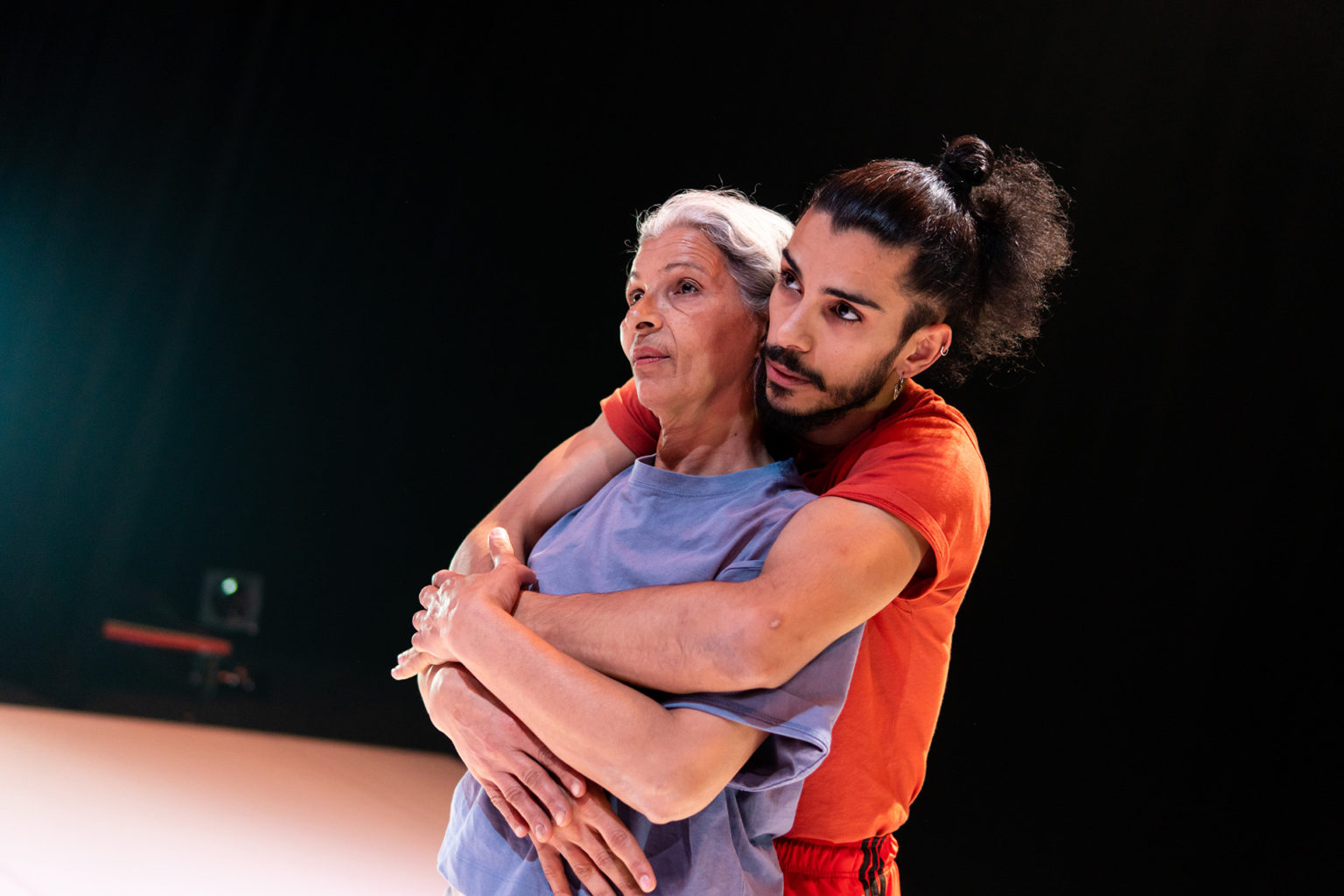 Le danseur Mohamed Toukabri est sur scène avec sa maman qu'il tient dans ses bras. il porte un t-shirt de couleur orange et sa maman prte un t-shirt de couleur mauve.