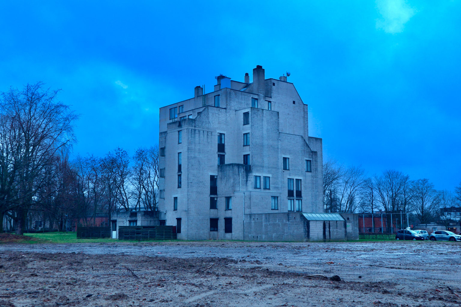 Photo représentant un immeuble, tout en béton, au bout d'un terrain en friche.