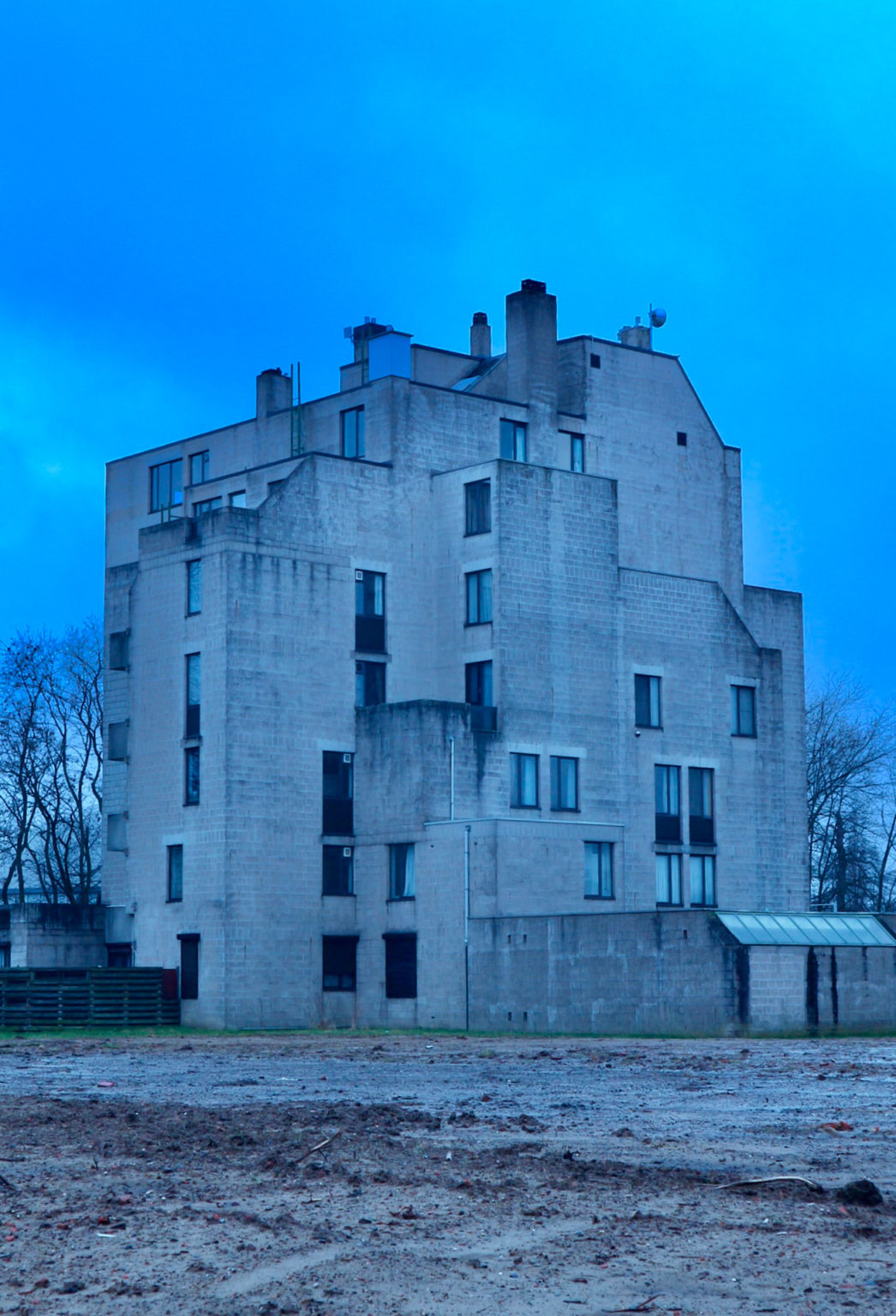 Photo représentant un immeuble, tout en béton, au bout d'un terrain en friche.