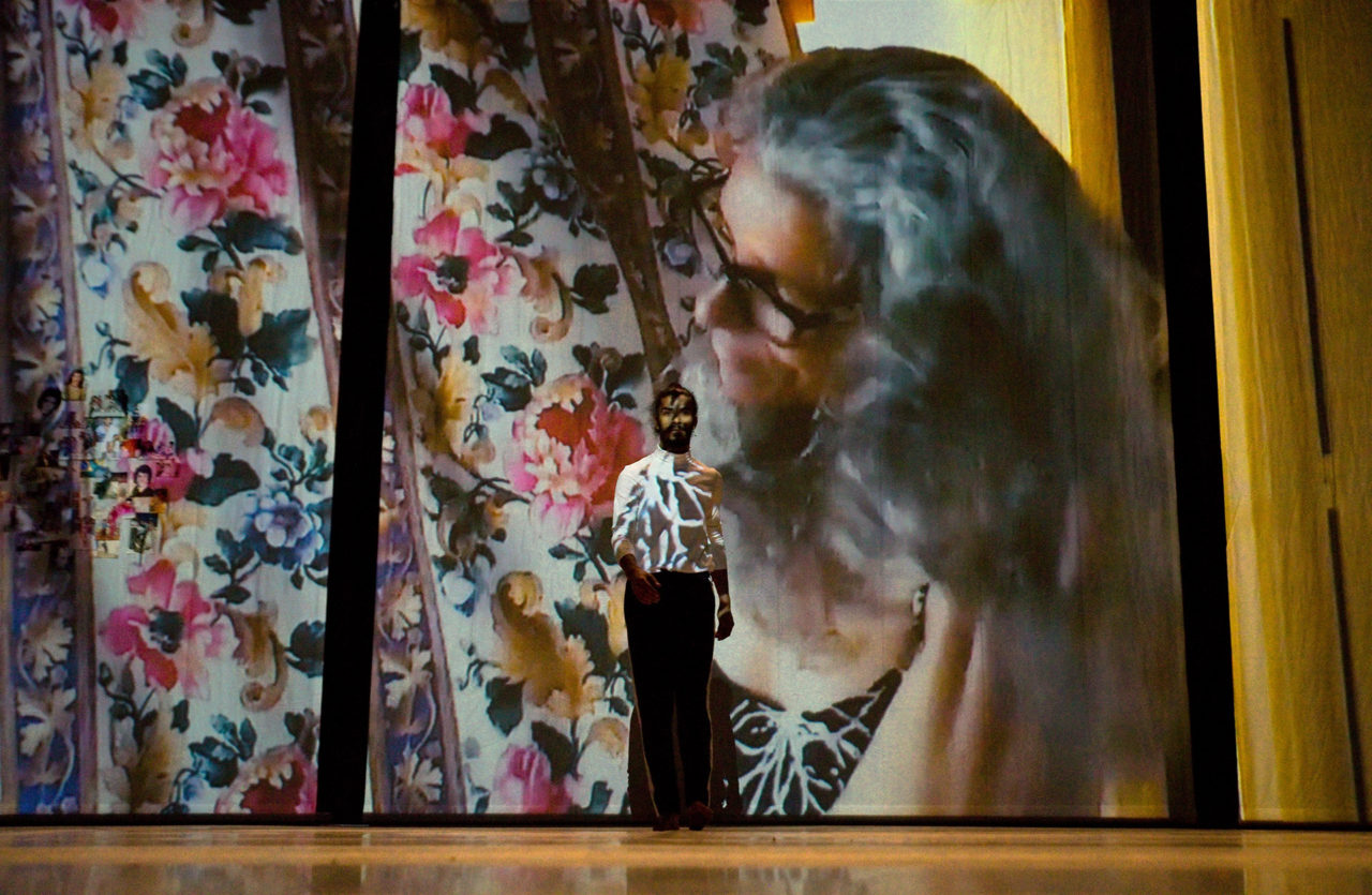 Photo du spectacle "The Upside Down Man" où m'on voit Mohamed Toubari sur une scène vide. Le portrait de sa mère derrière un rideau se reflète sur lui et sur un écran géant derrière lui.