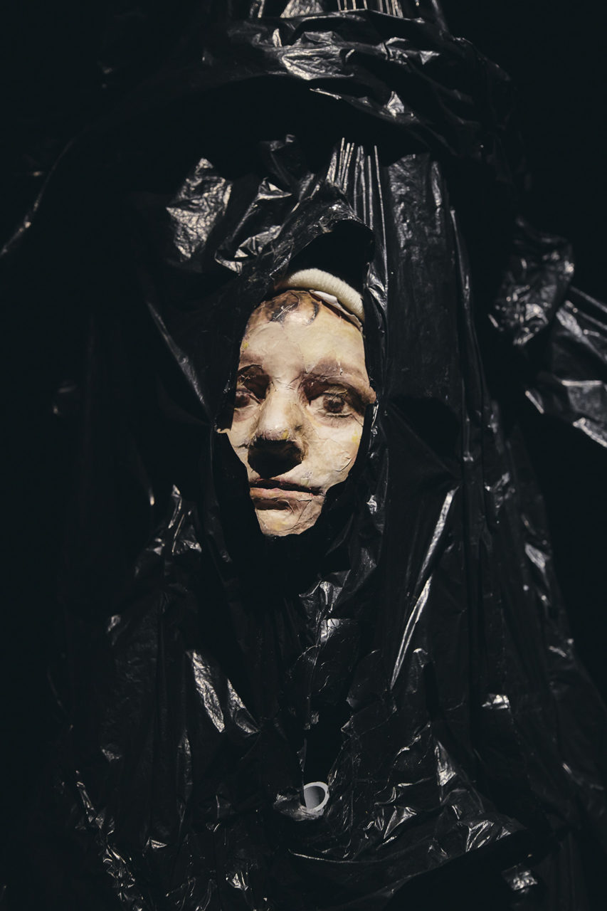 Une marionnette représentant un enfant est dans un sac-poubelle noir. Seul son visage en ressort.
