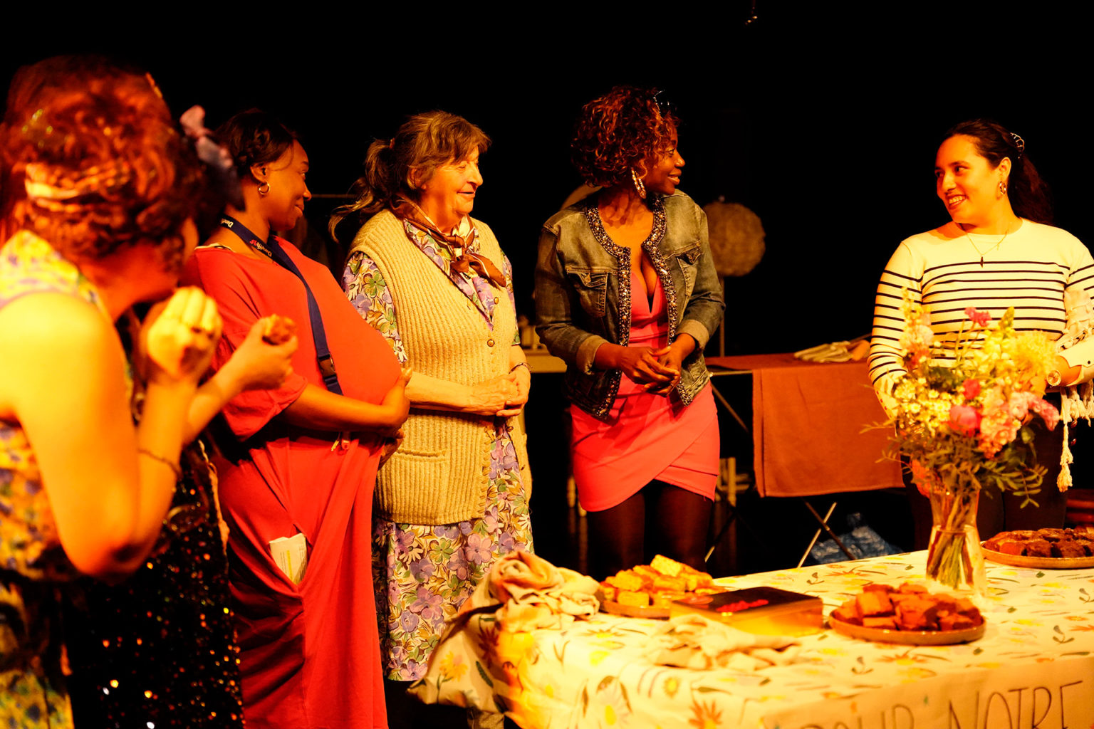 Participant·es du projet L'Atelier des femmes lors d'une représentation de "Habiter". Elles se regardent en souriant devant une table où il y a de quoi manger et un bouquet de fleurs.