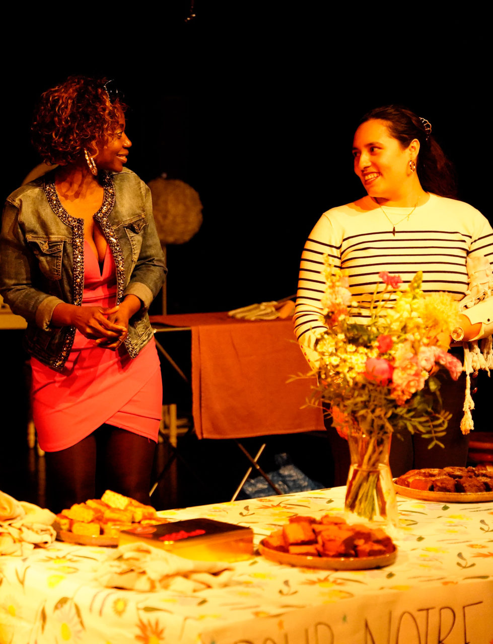 Participant·es du projet L'Atelier des femmes lors d'une représentation de "Habiter". Elles se regardent en souriant devant une table où il y a de quoi manger et un bouquet de fleurs.