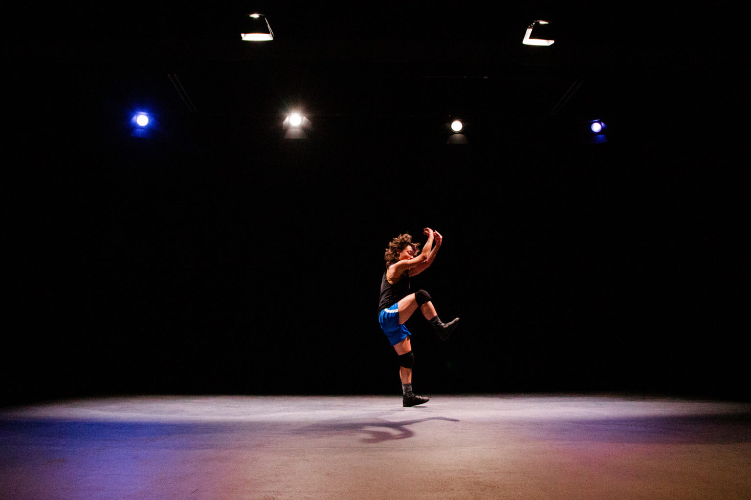 Une danseuse, en short, t-shirt et jambières, seule au milieu d'un plateau nu, danse et saute.