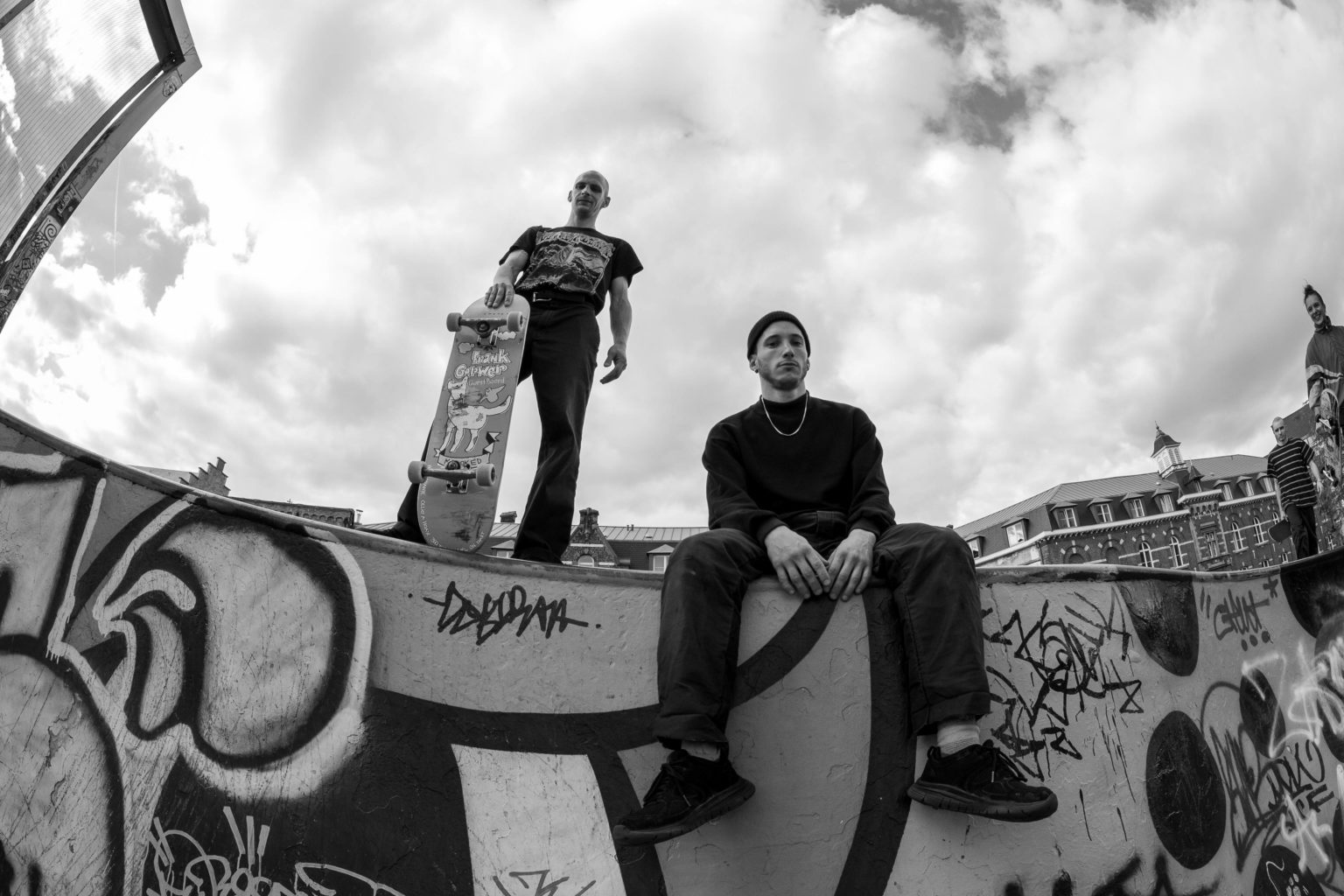 Un homme se tient assis sur le côté d'un skatepark. Un autre se tient debout à côté de lui avec un skateboard à la main.