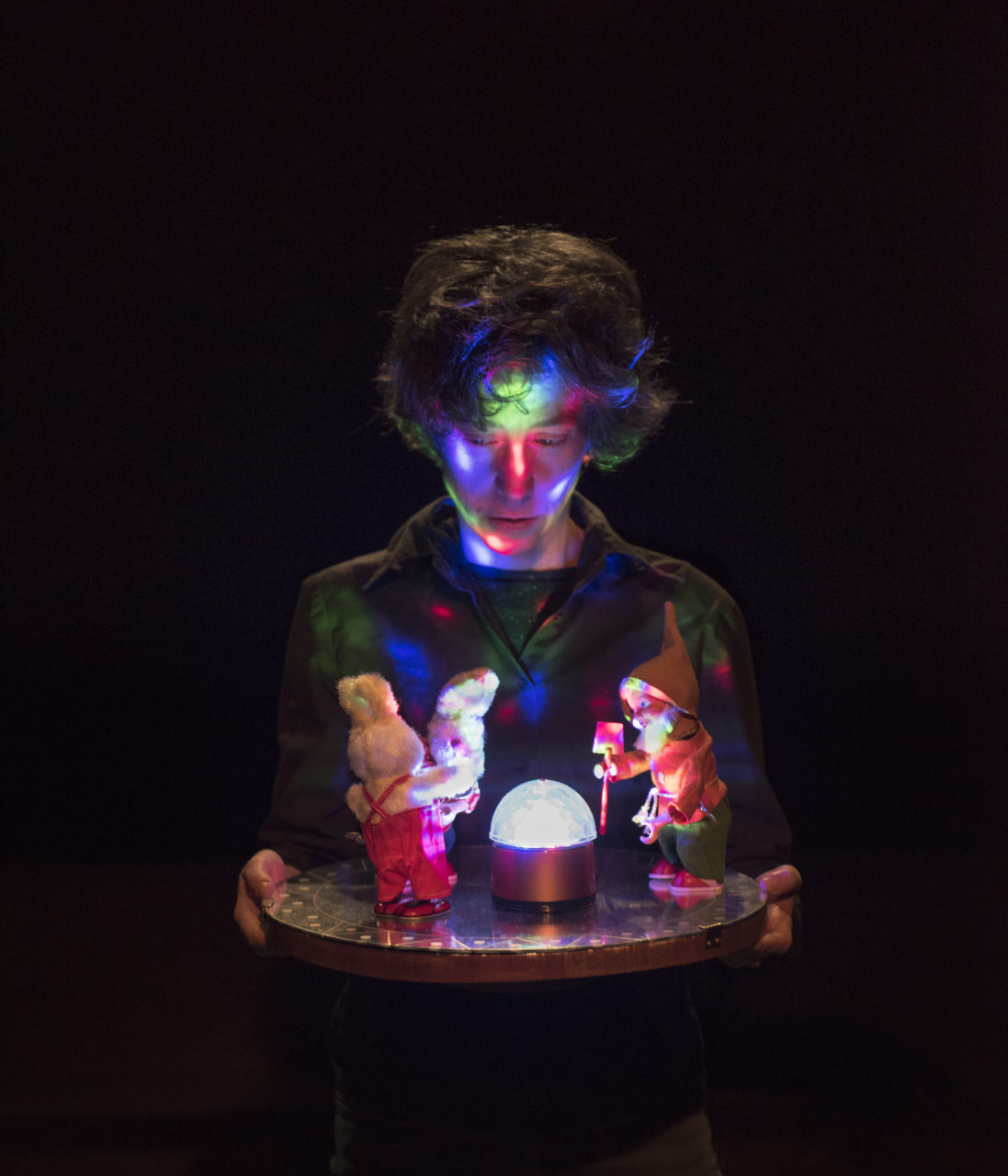 Un acteur tient des figurines sur un plateau avec une lumière multicolore qui le recouvre.