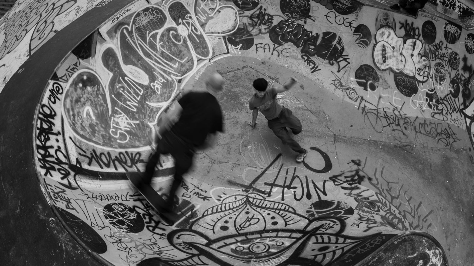 Dans un skatepark, un jeune homme fait du skate et un autre du breakdance.