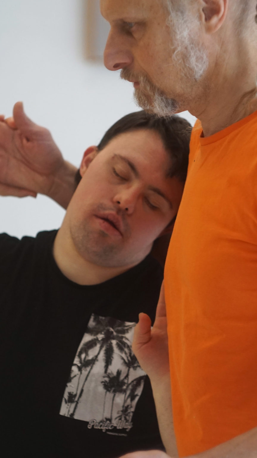 Un personne avec un t-shirt noir, les yeux fermés, repose sa tête sur la poitrine d'un autre homme avec un t-shirt orange et des cheveux gris. Ils sont en mouvement ensemble.
