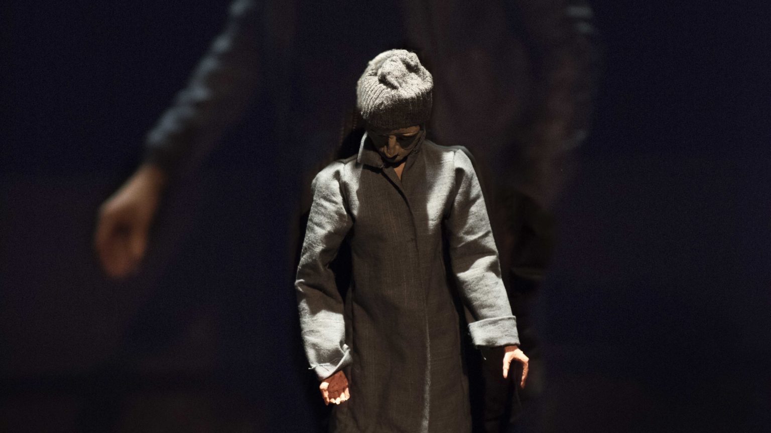 Une figurine habillée d'un manteau, d'une béret et de lunettes de soleil se tient devanat la caméra avec un acteur flouté qui se tient derrière elle.