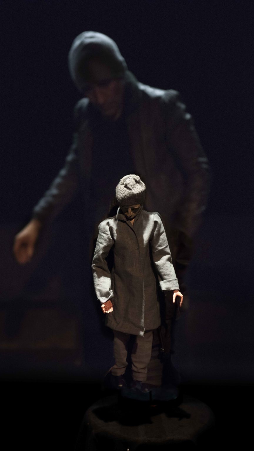 Une figurine habillée d'un manteau, d'une béret et de lunettes de soleil se tient devanat la caméra avec un acteur flouté qui se tient derrière elle.