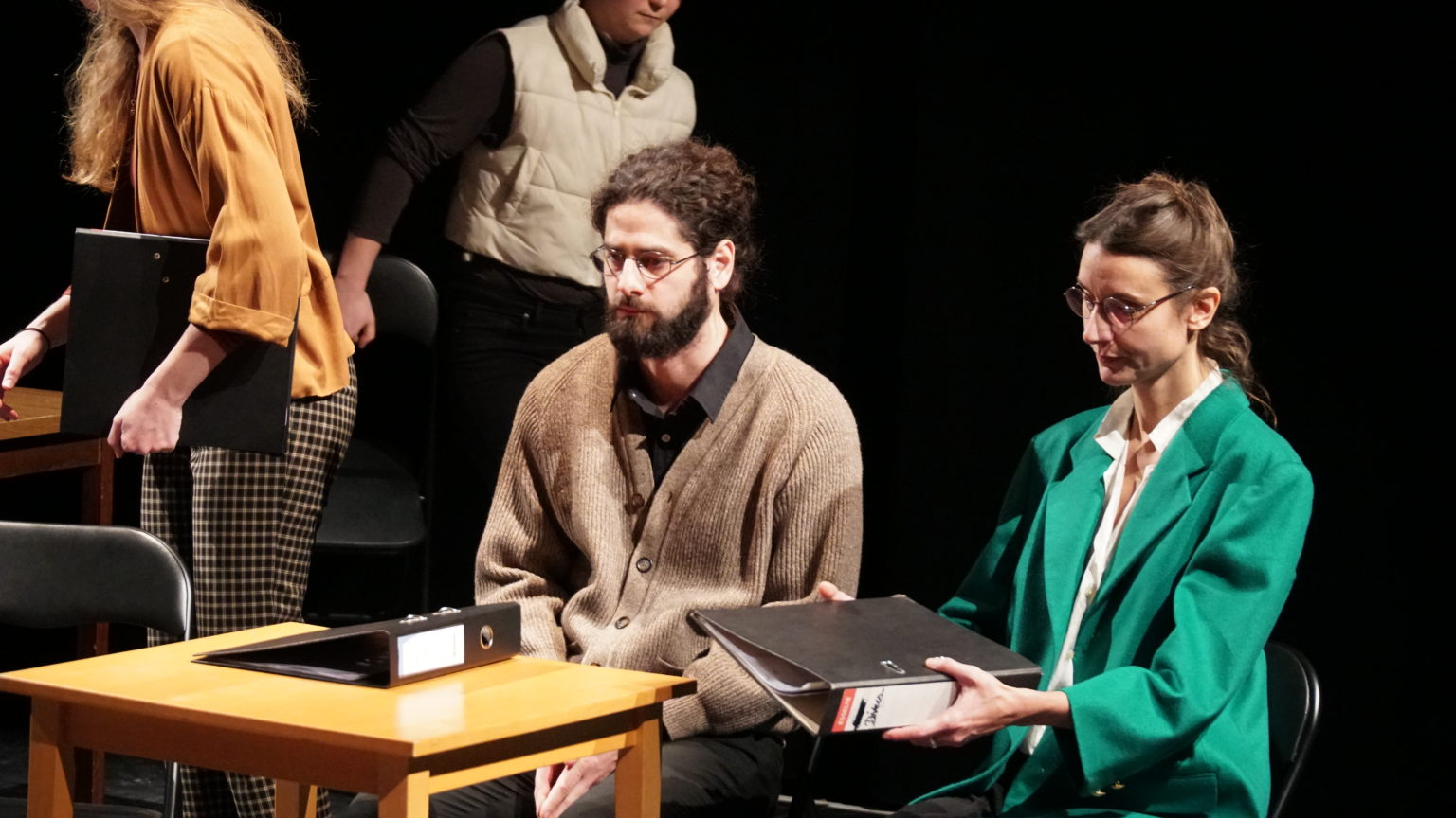 Un homme et une femme sont assis à une table avec un classeur posé dessus. La femme porte un blazer vert et des lunettes. Elle a les cheveux attachés et elle tient un classeur en main. L'homme porte des lunettes,  une chemise noire et un cardigan marron. Il regarde le vide. Des gens sont en train de s'installer sur des chaises derrière elleux.