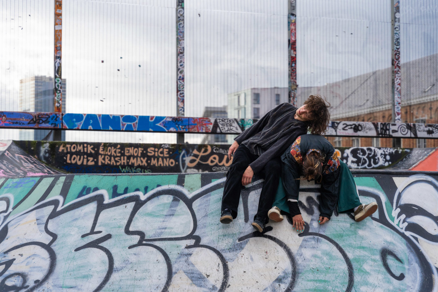 Un personne est assise pliée en 2 sur le côté d'un skatepark avec une autre personne assise à côté et appuyée sur elle.