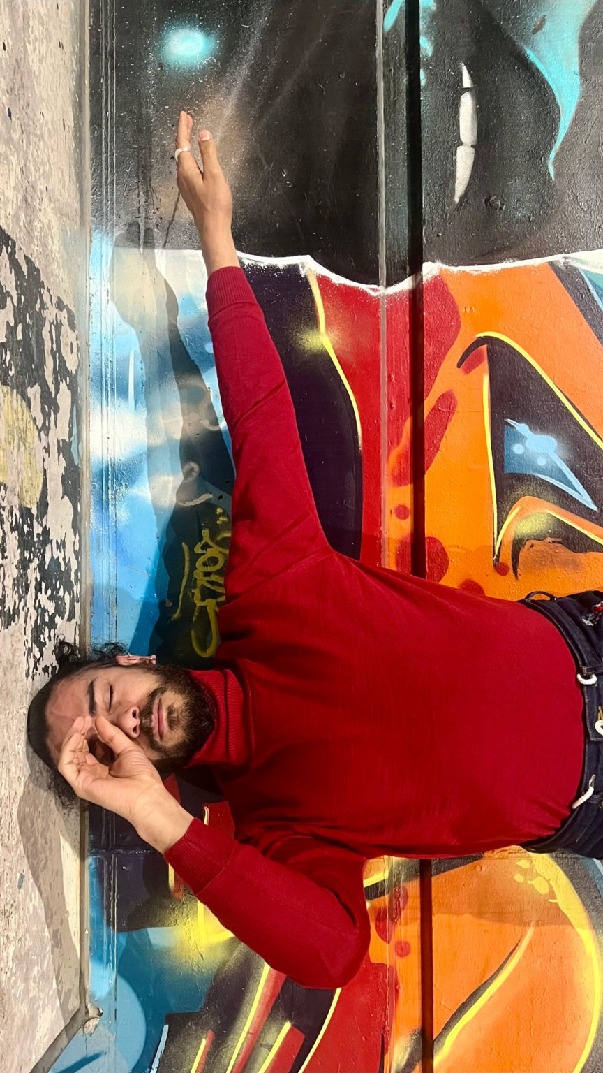 Mohamed Toukabri se tient devant un mur rempli de graffitis avec le bras tendu vers la droite et la main gauche en roind devant son oeil gauche. Il porte un pull rouge et à les cheveux attachés.