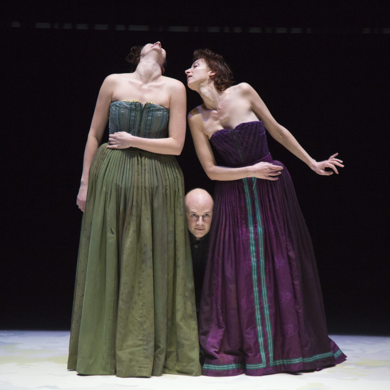 Deux femmes se tiennent debout sur scène avec un homme qui passe sa tête entre les deux avec la tête au niveau de leur hanche. La femme de gauche porte une robe bustier verte et se tient la tête en l'air. La femme de droite porte une robe bustier mauve et se tient la tête vers la femme de gauche.