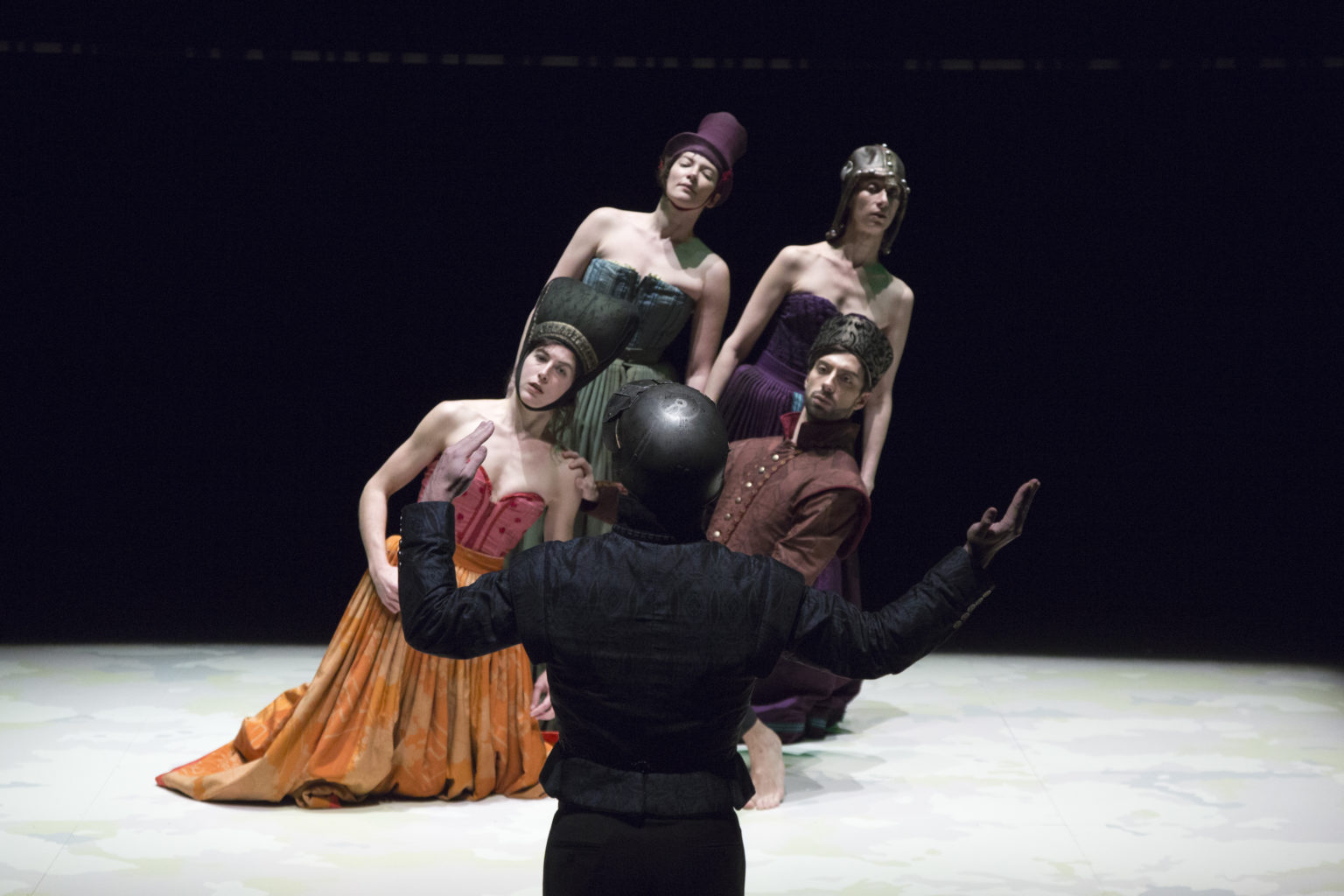 Quatre danseurs·euses se tiennent sur scène penchés vers la droite avec un homme face à elleux qui tient ses bras parallèles à son corps.