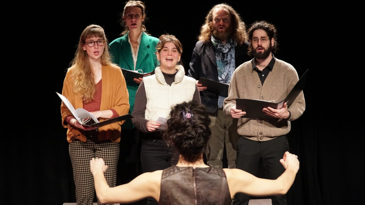 Une chorale chant en chœur. On voit une cheffer d'orchestre qui les mène de dos.