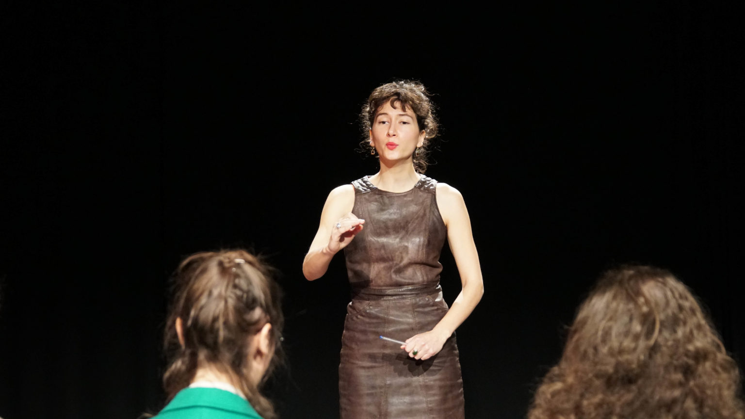 Un cheffe d'orchestre se tient debout une main levée en train de mener une chorale. Elle porte une robe marron et elle à des cheveux bouclés bruns attachés en chignon.