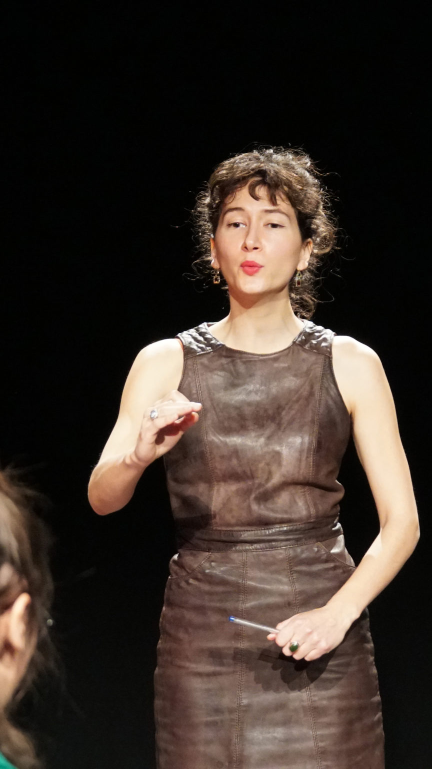 Un cheffe d'orchestre se tient debout une main levée en train de mener une chorale. Elle porte une robe marron et elle à des cheveux bouclés bruns attachés en chignon.