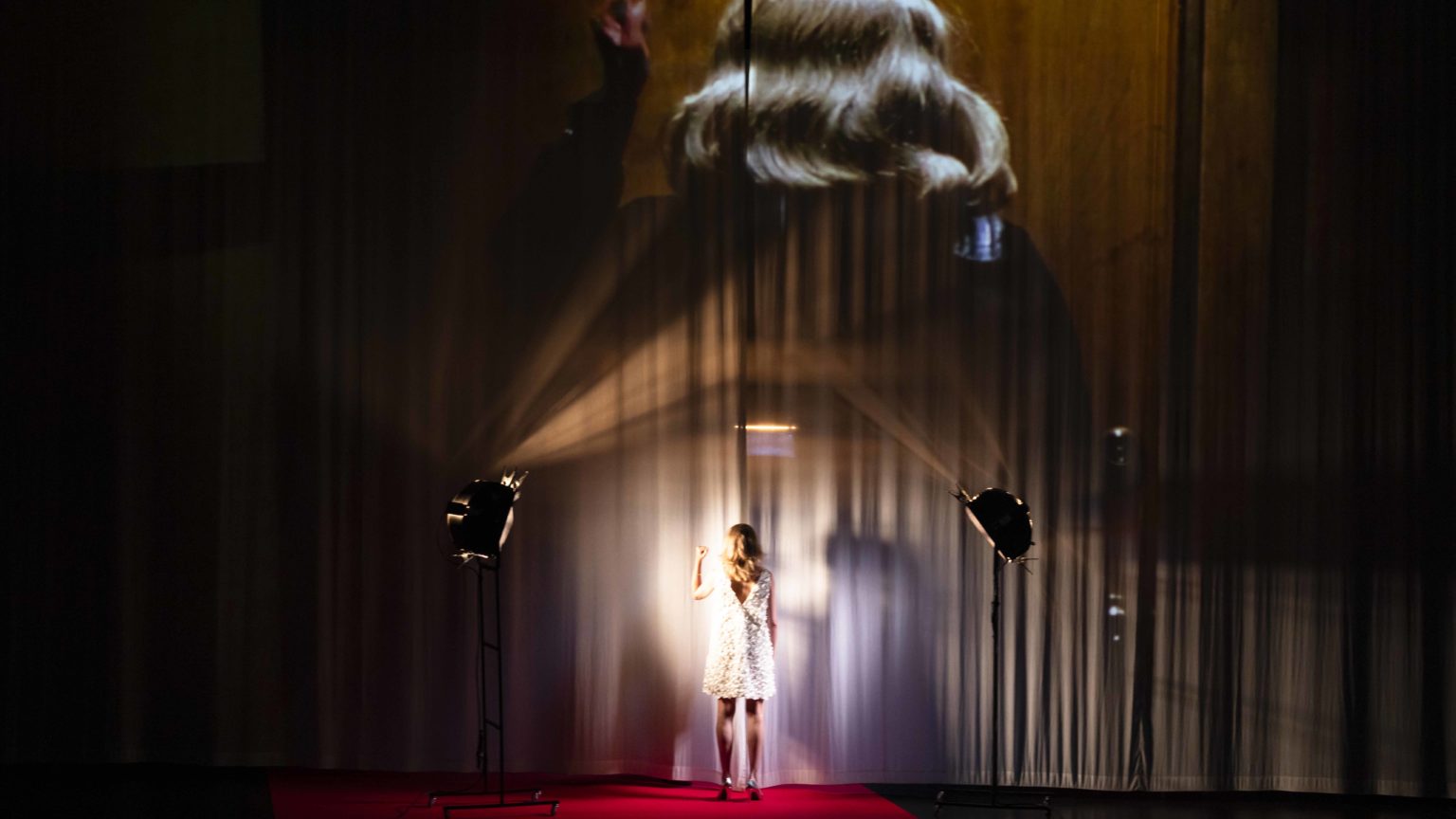 Une actrice de dos dans une robe blanche. Un rideau derrière elle sur lequel est projeté une vidéo.