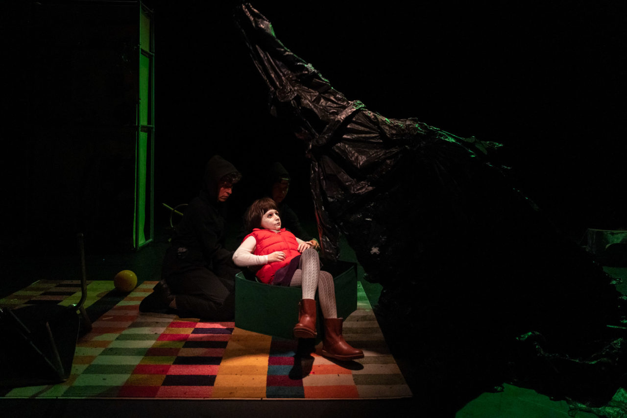 Une marionnette représentant une petite fille avec un gilet sans manches rouge. Elle est assise, près d'un grand objet, comme un arbre.