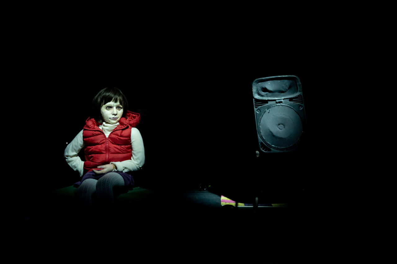 Une marionnette représentant une petite fille avec un gilet sans manches rouge. Un bafle se trouve à côté d'elle. Elle est assise.