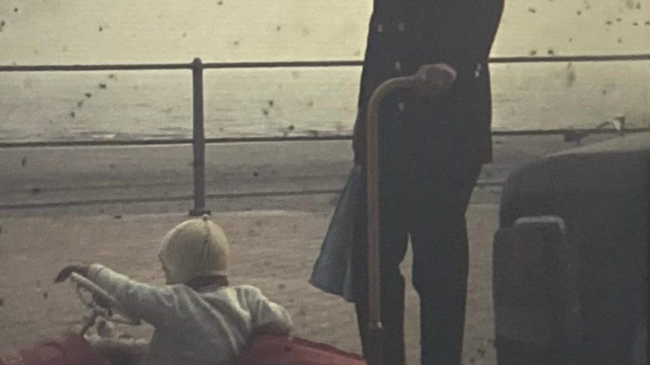 Photo vintage d'une personne qui tient une canne et regarde la caméra. Cette personne se trouve à côté d'un enfant dans une petite voiture rouge.
