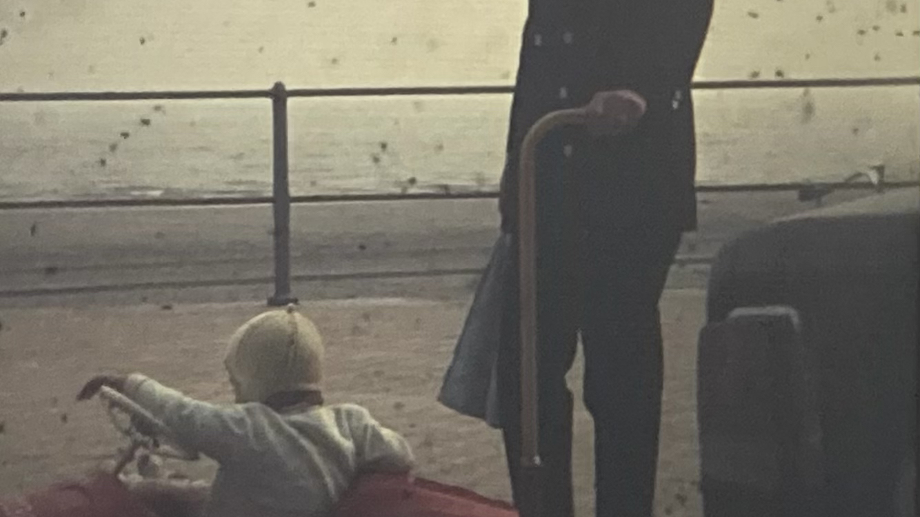 Photo vintage d'une personne qui tient une canne et regarde la caméra. Cette personne se trouve à côté d'un enfant dans une petite voiture rouge.