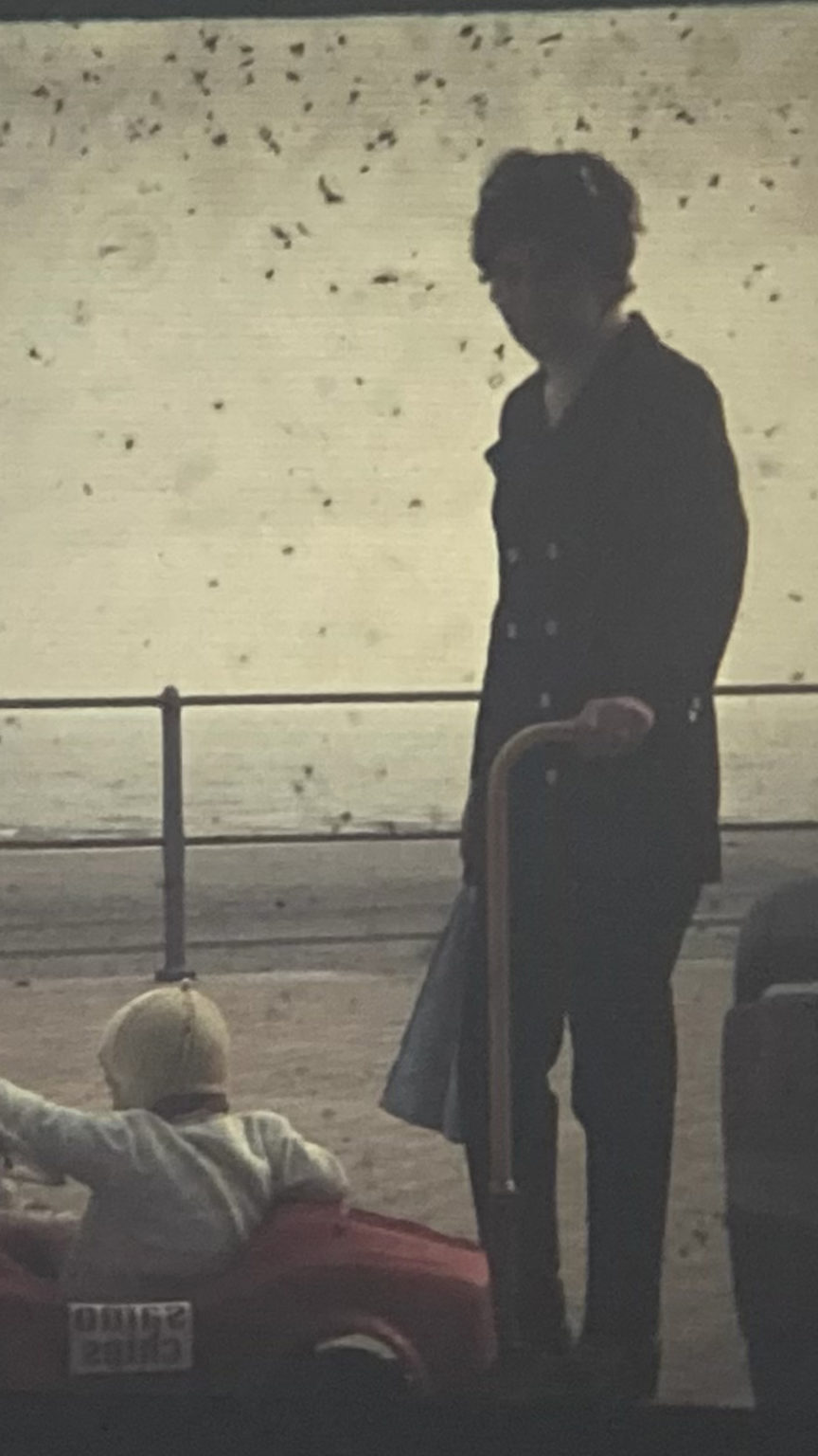 Photo vintage d'une personne qui tient une canne et regarde la caméra. Cette personne se trouve à côté d'un enfant dans une petite voiture rouge.
