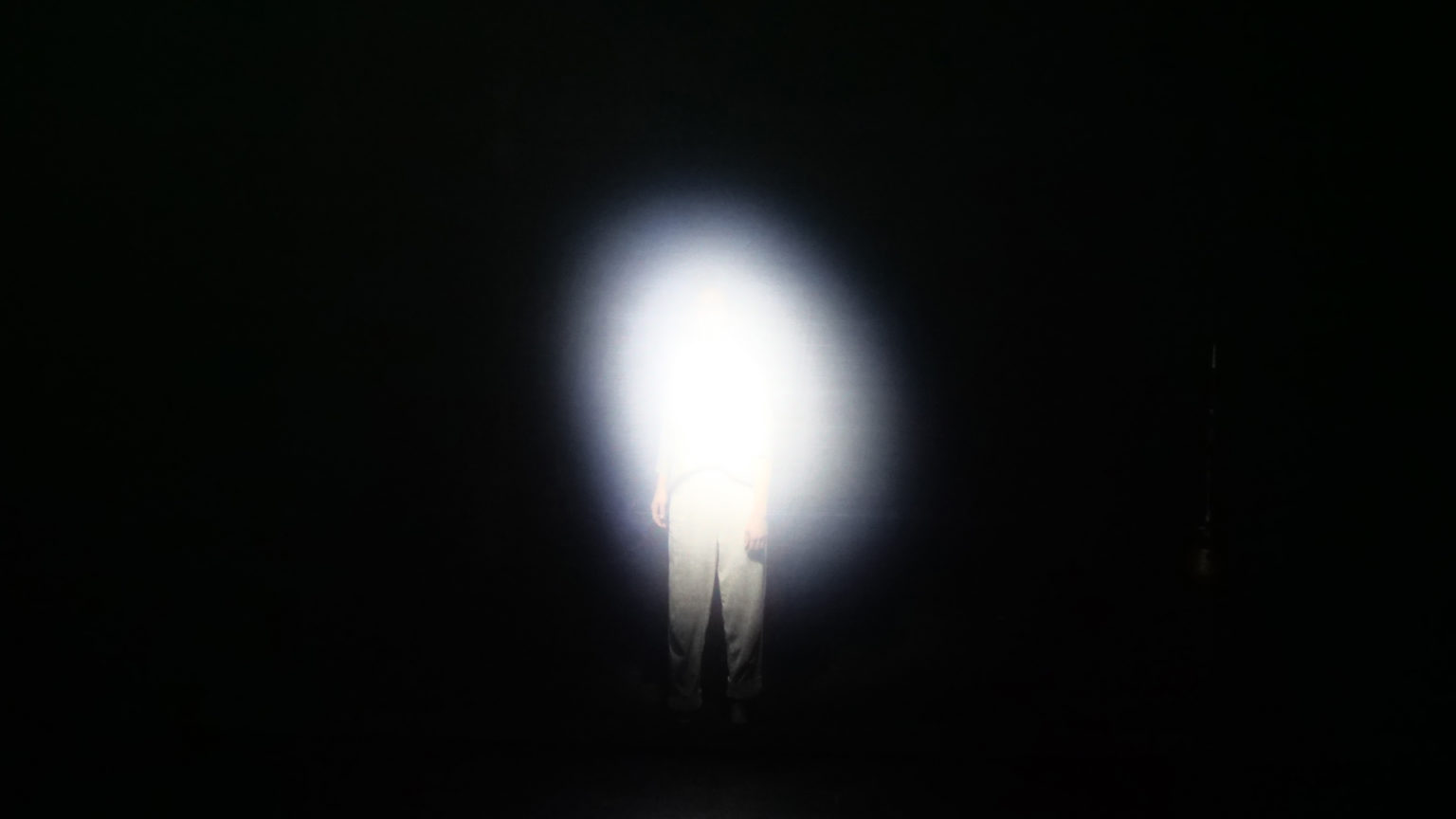 Une personne se tient dans le noir avec une lumière blanche qui cache une grande partie du haut de son corps.