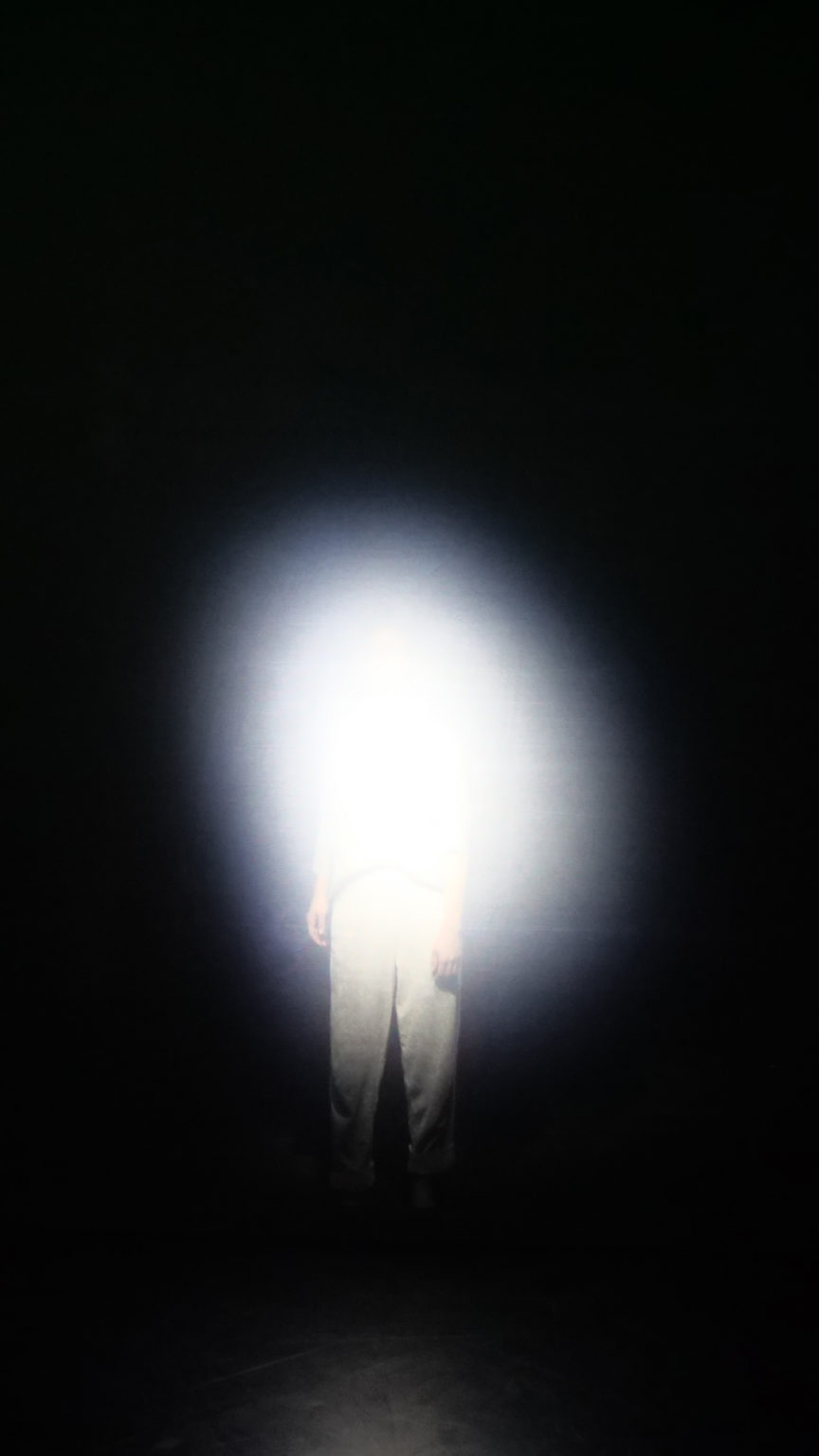 Une personne se tient dans le noir avec une lumière blanche qui cache une grande partie du haut de son corps.